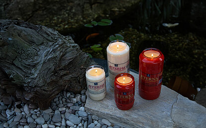 Zwei große und zwei kleine rote und weisse brennende Grabkerzen aus 100% Pflanzenöl. Zwei sind weiß, zwei rot. Die Grablichter stehen auf einer Steinplatte vor einem kleinen Teich.