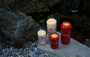 Zwei große und zwei kleine rote und weisse brennende Grabkerzen aus 100% Pflanzenöl. Zwei sind weiß, zwei rot. Die Grablichter stehen auf einer Steinplatte vor einem kleinen Teich.