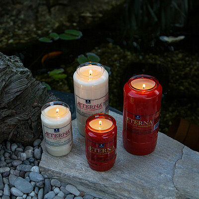 Zwei große und zwei kleine rote und weisse brennende Grabkerzen aus 100% Pflanzenöl. Zwei sind weiß, zwei rot. Die Grablichter stehen auf einer Steinplatte vor einem kleinen Teich.