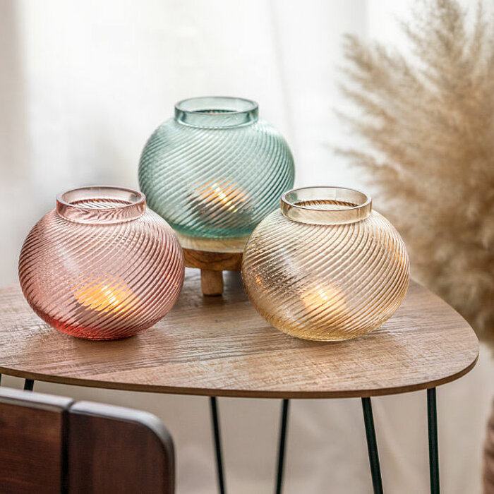 Drei "Sandra Rich" Kerzengläser in Rot, Grün und Gelb stehen auf einem hölzernen Beistelltisch. In den Gläsern brennen kleine Kerzen, die ein warmes Licht ausstrahlen. Im Hintergrund sind beige Vorhänge und eine Vase mit getrocknetem Pampasgras zu sehen.