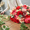 Ein opulenter Blumenstrauß, bestehend aus roten, rosa und weißen Rosen sowie einigen pinken Pfingstrosen, liegt auf einem Holztisch. Im Hintergrund arbeitet eine Person an weiteren Blumengestecken und arrangiert frische Blumen. Die Atmosphäre ist ruhig un