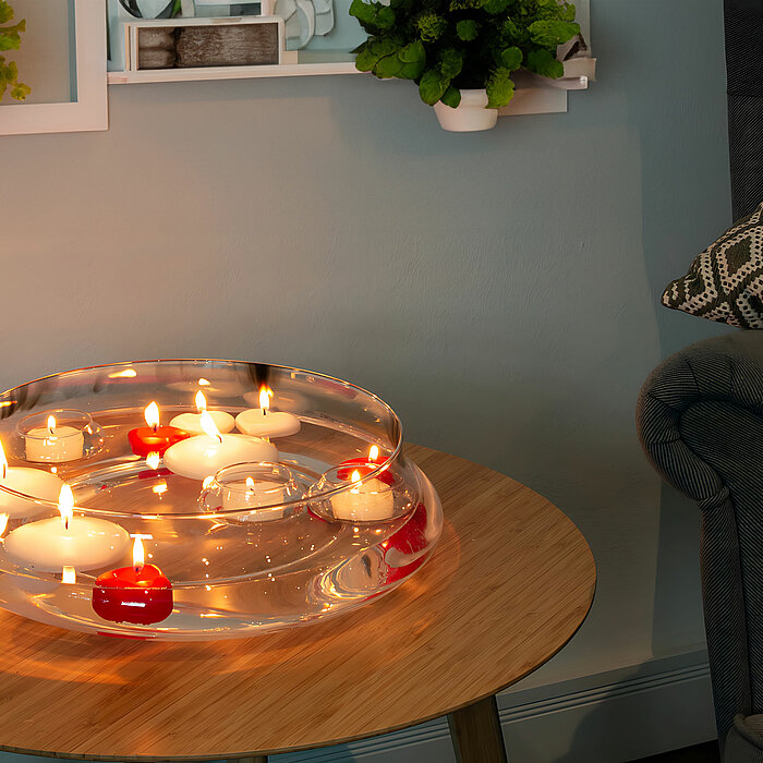 Auf einem runden, hölzernen Beistelltisch steht eine transparente Schale mit mehreren brennenden Kerzen von DecoLite, darunter rote und weiße. Im Hintergrund befinden sich grüne Pflanzen auf Wandregalen und ein dunkelgrauer Sessel mit einem gemusterten Ki