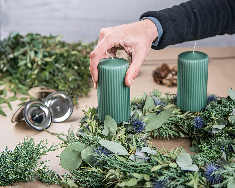Eine menschliche Hand hält eine grüne geriffelte Stumpenkerze und stellt sie auf einen Kranz, der mit Zweigen und Blättern umgeben ist. Außen herum liegen weitere Zweige und einige Kerzenhalter mit Dorn.