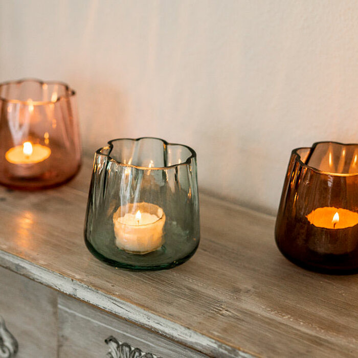 Drei brennende Teelichter in farbigen, wellenförmigen Glasbehältern von Sandra Rich stehen auf einer antiken, verzierten Kommode. Die Glasbehälter sind in den Farben Rosa, Grün und Braun gehalten. Rechts daneben steht eine weiße, brennende Stumpenkerze. I