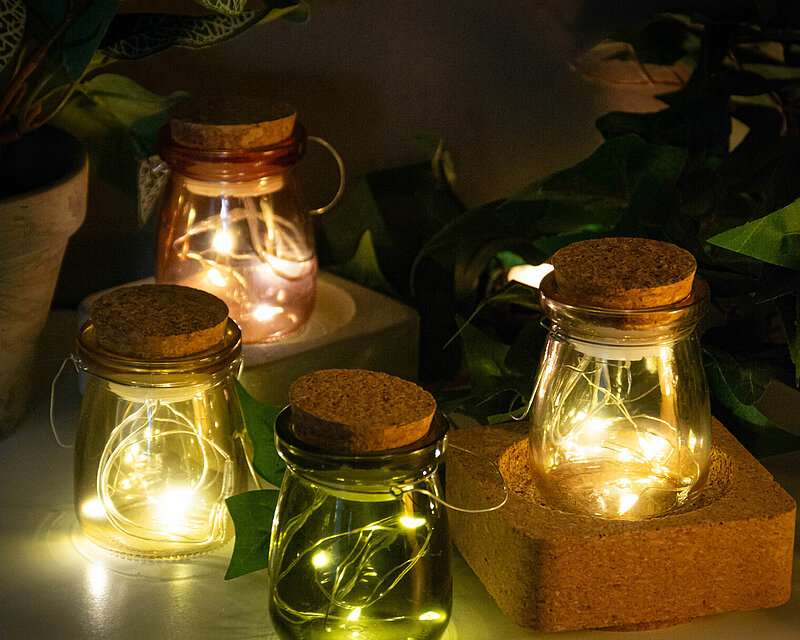 Das gesamte Bild hat eine recht dunkle, aber schöne und gemütliche Ausstrahlung. Auf einem weißen Tisch stehen vier unterschiedlich farbene LED-Krüge von Edelman, jeweils mit einer brennenden Mikro-LED-Kette darin. Die warmen Lichter der LED-Krüge strahlen sanft und schaffen eine beruhigende, einladende Atmosphäre. Hinter den Krügen steht eine üppige Topfpflanze mit sattem Grün, und einige Zweige grünen Efeus ranken sich elegant um das Arrangement. 