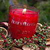 Eine brennende rote Kerze vom Hersteller "Prices Candles" mit der Aufschrift "For Santa" steht in einem Glas auf einer dekorativen Unterlage aus getrockneten Früchten und grünen Zweigen mit roten Beeren. Im Hintergrund sind verschwommene grüne Tannenzweig