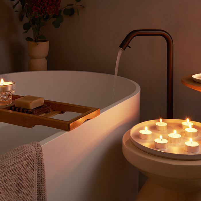 Mehrere brennende Bolsius Teelichter verteilen warmes, atmosphärisches Licht im Raum. Einige Teelichter stehen auf einem Holztablett, das quer über einer gefüllten Badewanne mit einem Buch, Seife und einem Glas platziert ist. Weitere brennende Bolsius Tee