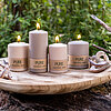 Vier brennende Kerzen des Herstellers Wenzel, in einer neutralen Beige-Farbe, stehen auf einem runden Holztablett. Die Kerzen haben verschiedene Größen und tragen die Aufschrift "PURE Natural Wax". Das Arrangement steht auf einem Baumstamm oder einer Stei