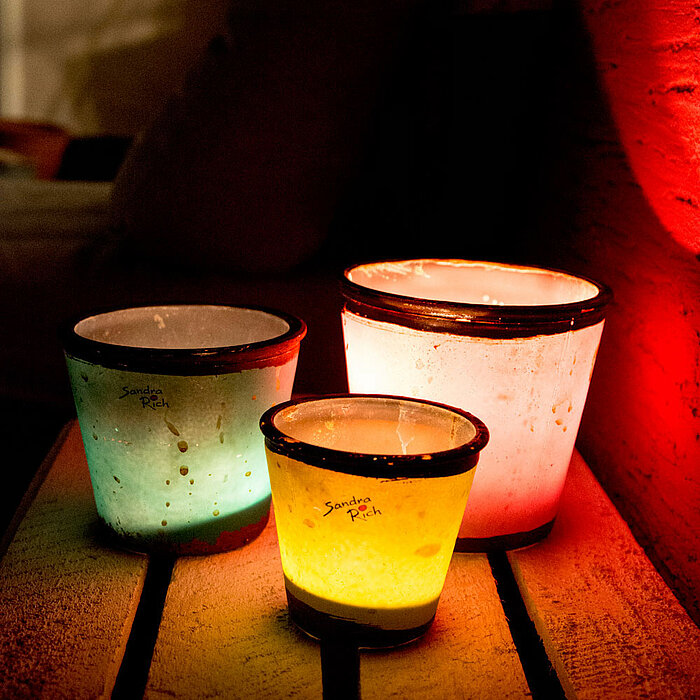 Drei farbige Teelichtgläser vom Hersteller "Sandra Rich" stehen auf einer Holzpalette. Die Gläser sind in Grün, Gelb und Rot beleuchtet und verleihen der Umgebung eine warme und gemütliche Atmosphäre.