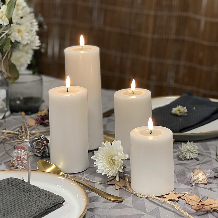 Vier brennende weiße Stumpenkerzen in verschiedenen Größen auf einem festlich gedeckten Tisch. Die Kerzen sind umgeben von getrockneten Blättern und Blumen. Auf dem Tisch liegen zwei Teller mit dunkelgrauen Servietten, die von kleinen Blumendekorationen g