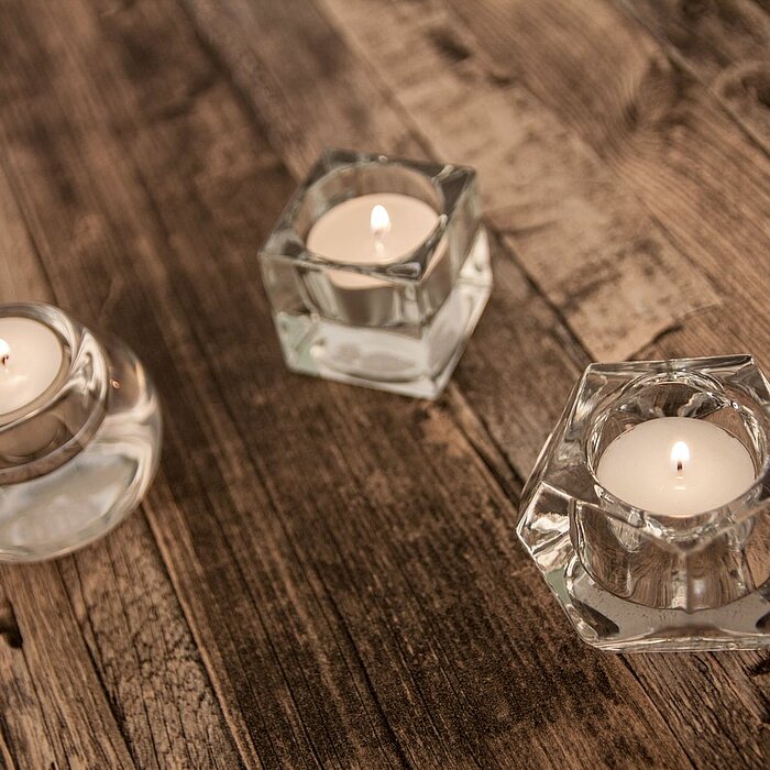 Zwei brennende Teelichter in transparenten Glasbehältern vom Hersteller Sandra Rich. Das eine Teelicht befindet sich in einem runden Glasgefäß, das andere in einem facettierten, kristallartigen Glas, beide auf einer rustikalen Holzoberfläche platziert.