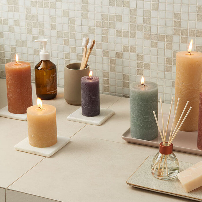 Sechs brennende Bolsius Rustikkerzen in verschiedenen Größen und Farben (Beige, Braun, Anthrazit, Grün und Orange) stehen auf Untersetzern und Tabletts in einem Badezimmer. Die Kerzen sind auf einem beige gefliesten Waschtisch verteilt. Zudem befinden sic