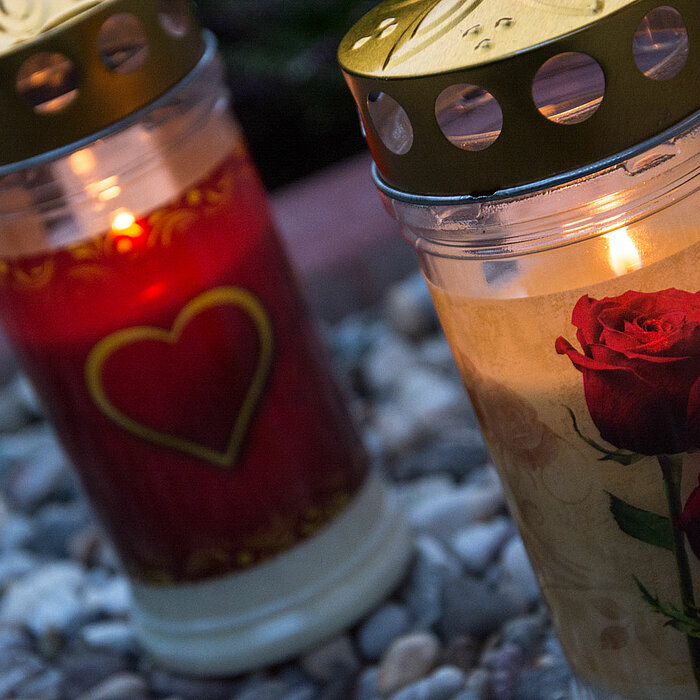 Das Bild zeigt zwei leidenschaftlich brennende Grablampen auf einem kiesigen Boden. Die linke Kerze ist rot mit einem goldenen Herzmotiv, während die rechte Kerze ein Bild von zwei roten Rosen auf beigem Hintergrund zeigt. Beide Kerzen sind mit goldenen D