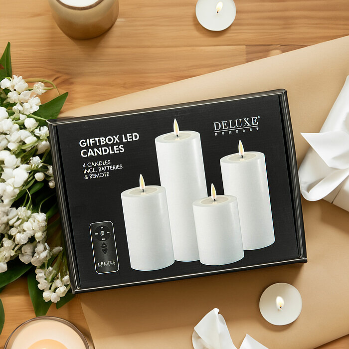 Vier brennende weiße Stumpenkerzen in verschiedenen Größen auf einem festlich gedeckten Tisch. Die Kerzen sind umgeben von getrockneten Blättern und Blumen. Auf dem Tisch liegen zwei Teller mit dunkelgrauen Servietten, die von kleinen Blumendekorationen g