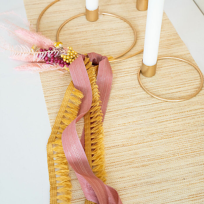 Vier weiße Kerzen in goldenen Kerzenhaltern, die auf einem hellen Tischläufer stehen. Um die Kerzen sind dekorative Bänder in Rosa und Gelb geflochten, verziert mit rosa und gelben Trockenblumen. Die Produkte sind vom Hersteller "Halbach".