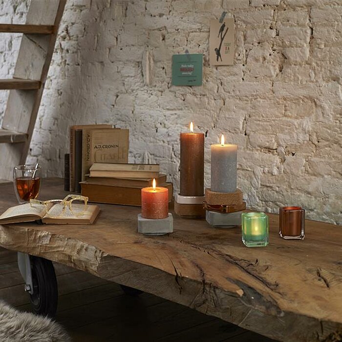 Drei brennende Stumpenkerzen in verschiedenen Farbtönen und Größen stehen auf einem rustikalen Holztisch vor einer weiß gestrichenen Backsteinwand. Eine der Kerzen ist grün, eine braun und die dritte orange. Auf dem Tisch befinden sich ein aufgeschlagenes