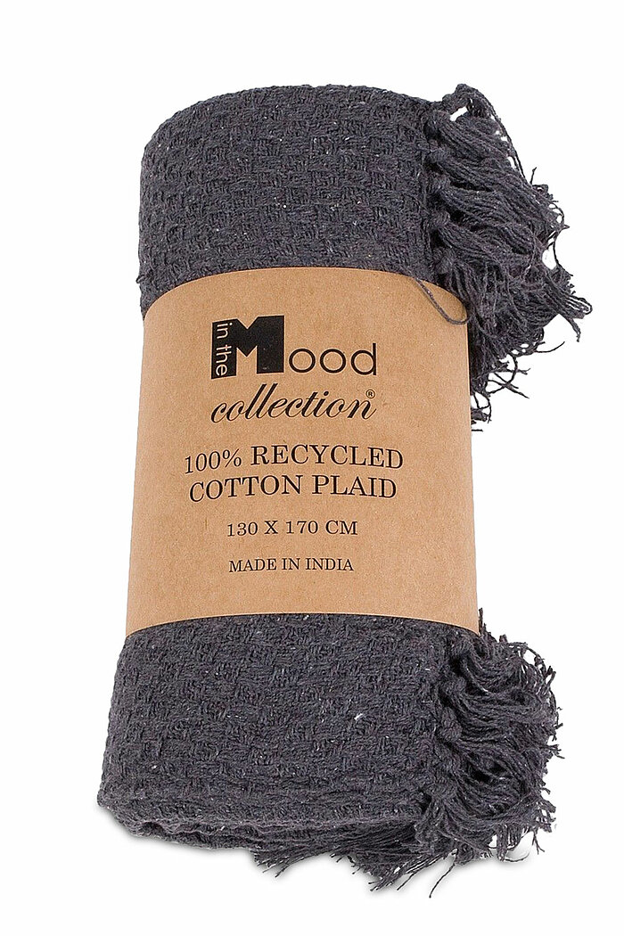 Eine dunkelgraue Decke, zu einem kompakten Bündel gerollt und mit einer braunen Papierbanderole umwickelt. Auf der Banderole steht in schwarzer Schrift: "Edelman" und "100% RECYCLED COTTON PLAID", darunter in etwas kleinerer Schrift "130 X 170 CM" und "MA