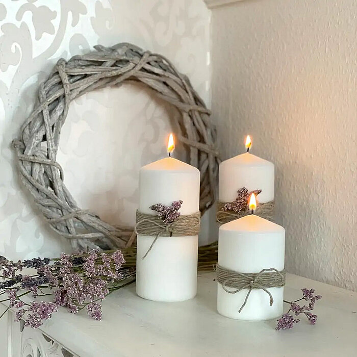 Drei brennende Müller Stumpenkerzen in Weiß, dekoriert mit Juteschnur und violetten Blüten, stehen auf einer weißen Oberfläche. Im Hintergrund ist ein dekorativer Zweigkranz an der weißen Wand angebracht.