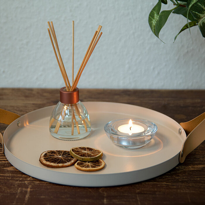 Ein Duftdiffusor von Edelman mit Holzstäbchen und eine brennende Teelicht-Kerze in einem Glasbehälter stehen auf einem runden Tablett mit seitlichen Lederschlaufen. Neben den Produkten von Edelman liegen getrocknete Orangenscheiben. Im Hintergrund ist ein
