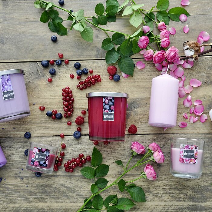 Verschiedene Müller Duftkerzen in Glasgefäßen und Stumpenkerzen in den Farben Lila, Rosa und Weiß sind auf einem Holztisch arrangiert. Die Kerzen sind von Blumen wie Lavendel, Rosen und Baumwollblüten sowie von Beeren wie Johannisbeeren und Blaubeeren umg