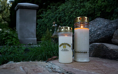 Ein kleines und ein großes weisses brennendes Grablicht mit einem Deckel stehen auf einer Steinplatte. vor einem großen Grabstein einer grünen Hecke und Gräsern.
