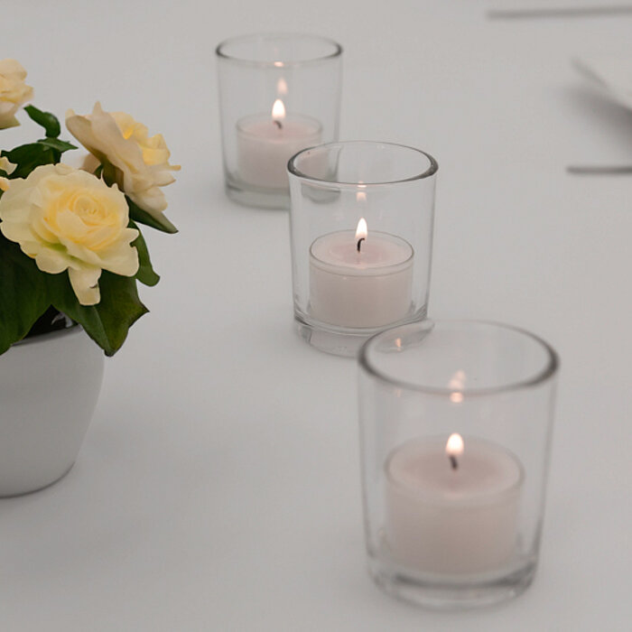 Zwei brennende weiße Kerzen in klaren Glasbehältern stehen auf einer Marmorplatte. Daneben liegt eine große rosafarbene Blume. Die Szene vermittelt eine ruhige und entspannende Atmosphäre.