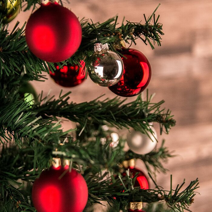 Grüne Zweige eines Weihnachtsbaums, geschmückt mit roten und silbernen Kugelornamenten. Der Baum fügt sich vor einem unscharfen, hölzernen Hintergrund, was eine gemütliche und festliche Atmosphäre schafft. Die roten Kugeln dominieren das Bild und vermitte