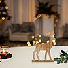 Ein gemütliches Wohnzimmer mit weihnachtlichem Flair. Im Vordergrund steht ein Tisch, auf dem ein Teelicht in einem kleinen Glaswindlicht, getrocknete Orangenscheiben und ein hölzerner Hirsch aufgestellt sind. Daneben liegen einige grüne Zweige. Im Hinter