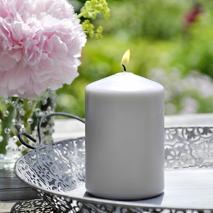 Drei brennende Müller Kerzen in grauen und pastellrosa Farbtönen stehen auf einem weißen Tisch. Es sind zwei Stumpenkerzen und eine Glaswindlichtkerze mit einem Blumenetikett des Herstellers "Müller". Rechts daneben befindet sich eine weitere kleine Glasw