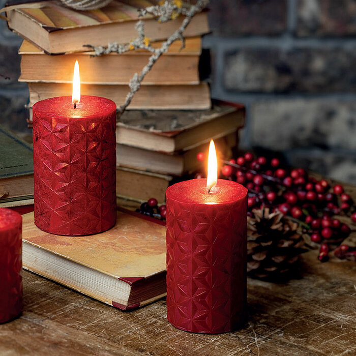 Drei brennende Wenzel Stumpenkerzen in verschiedenen Größen in einem tiefen Rotton stehen auf einem Holztisch vor einem Stapel Bücher. Die Kerzen sind mit einem geometrischen Muster verziert. Um die Kerzen herum sind Tannenzapfen und Zweige mit roten Beer