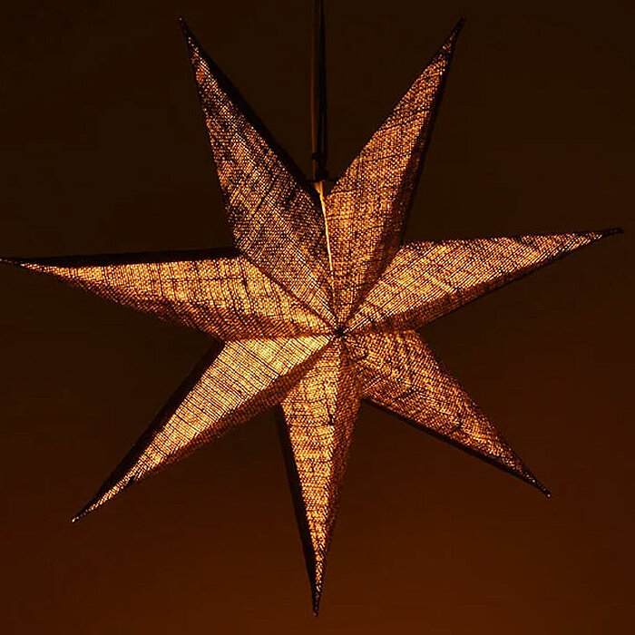 Ein leuchtender, siebenstrahliger Papierstern in warmen, goldenen Orangetönen hängt vor einem dunkelbraunen Hintergrund. Die feine Struktur des Papiers lässt das Licht weich durchscheinen und erzeugt eine beruhigende, warme Atmosphäre.