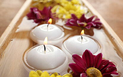 Drei brennende Schwimmkerzen treiben in einer Wasserschale zwischen pinken und gelben Blüten.