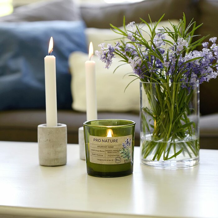 Zwei brennende weiße Stabkerzen in schlichten, grauen Haltern und eine grüne "Pro Nature" Duftkerze von Müller stehen auf einem weißen Tisch. Daneben befindet sich ein Glasgefäß mit einem Strauß lila Blumen. Im Hintergrund sind ein graues Sofa mit blauen 