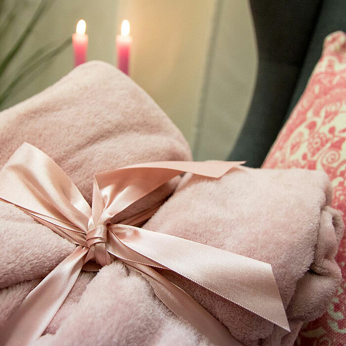 Zwei rosa Stabkerzen brennen im Hintergrund und tauchen die Szene in ein warmes Licht. Im Vordergrund liegt eine flauschige, altrosa Decke, die mit einem hellrosa Satinband zu einer Schleife gebunden ist. Rechts daneben liegt ein Kissen, das mit einem det