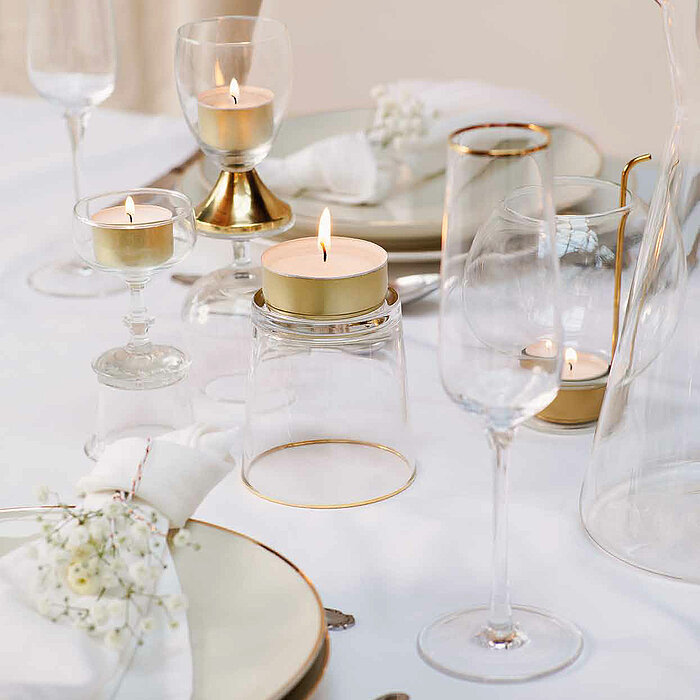 Ein eleganter, gedeckter Tisch mit einem minimalistischen und doch festlichen Ambiente. Drei brennende Bolsius-Teelichter in goldenen Haltern sind sorgfältig aufgestellt. Die Teelichter beleuchten stilvolle Champagnergläser, einen gläsernen Krug, Besteck 