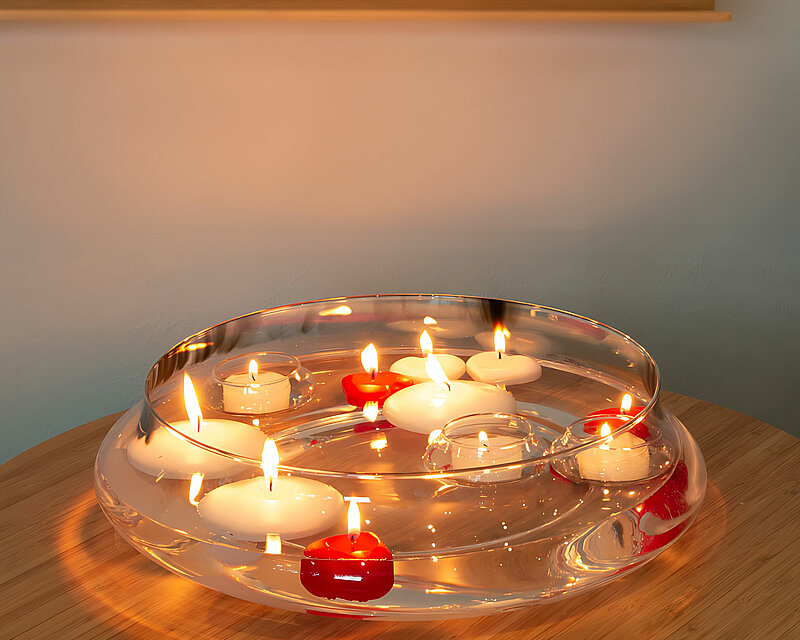 Im Hintergrund befindet sich ein Fenster. Auf dem Fensterbrett stehen drei Weiße Blumenkübel mit verschiedenen grünen Pflanzen. im Vordergrund steht ein runder Beistelltisch. Auf diesem runden Beistelltisch steht eine große Glasschale gefüllt mit Wasser und unterschiedlichen weißen und roten brennenden Schwimmkerzen.