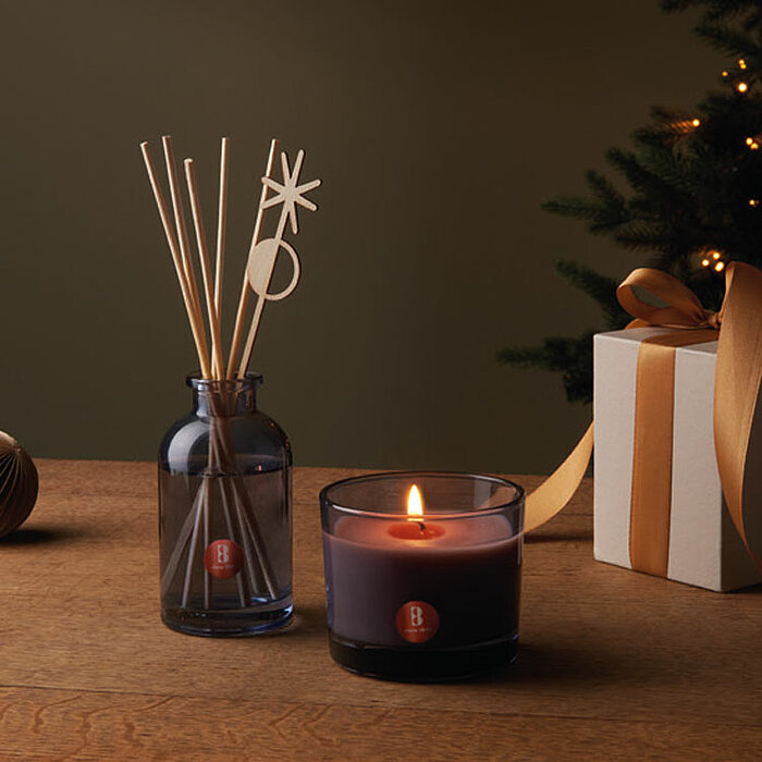 Eine brennende dunkelviolette Kerze in einem Glasbehälter steht auf einem Tisch. Daneben befindet sich eine dunkelblaue Glasflasche mit Duftstäbchen, die dekorative Holzaufsätze haben. Im Hintergrund ist teilweise ein geschmückter Weihnachtsbaum und ein f