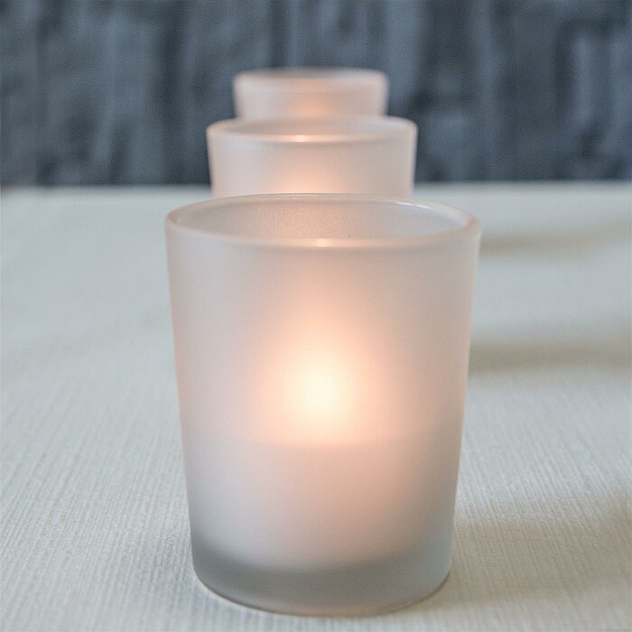 Zwei brennende Teelichter in matten Glasbehältern des Herstellers Sandra Rich stehen auf einer marmorierten Oberfläche. Beide Kerzen strahlen ein warmes, sanftes Licht aus.