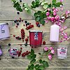 Verschiedene Müller Duftkerzen in Glasgefäßen und Stumpenkerzen in den Farben Lila, Rosa und Weiß sind auf einem Holztisch arrangiert. Die Kerzen sind von Blumen wie Lavendel, Rosen und Baumwollblüten sowie von Beeren wie Johannisbeeren und Blaubeeren umg