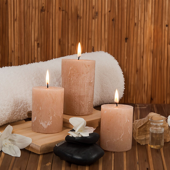 Zwei brennende rote Bolsius Rustikkerzen in verschiedenen Größen stehen nebeneinander auf einem Holztisch. Im Vordergrund sind Topfpflanzen mit bunten Blüten in Weiß, Gelb und Rot zu sehen. Im Hintergrund sind unscharfe grüne Pflanzen erkennbar.