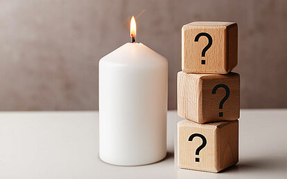 Auf einem Hellen Tisch befindet sich mittig in dem Bild eine weiße brennende Stumpenkerze. Rechts daneben sind drei aufeinander gestapelte Holzwürfel mit einem Fragezeichen auf der vorderen Seite.