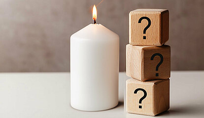 Auf einem Hellen Tisch befindet sich mittig in dem Bild eine weiße brennende Stumpenkerze. Rechts daneben sind drei aufeinander gestapelte Holzwürfel mit einem Fragezeichen auf der vorderen Seite.