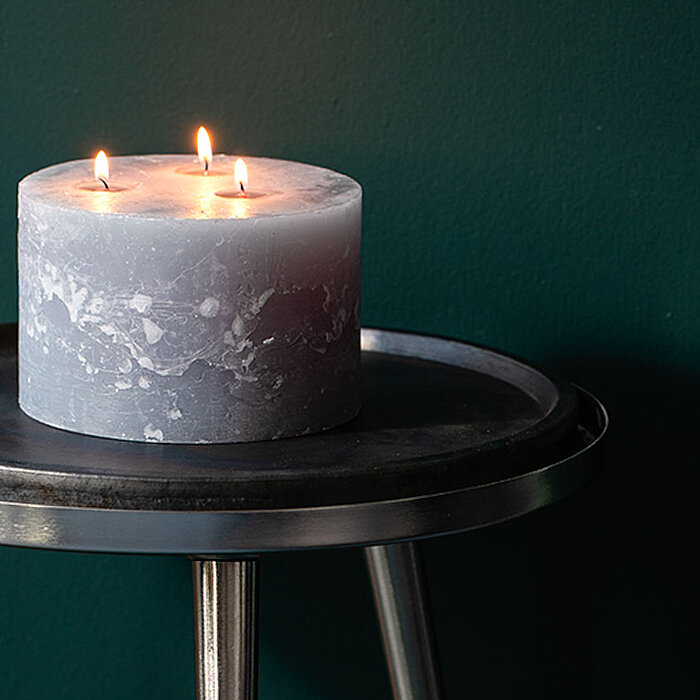 Eine brennende Bolsius Rustikkerze in Grau mit drei Flammen steht auf einem runden, metallischen Tisch vor einer dunkelgrünen Wand.