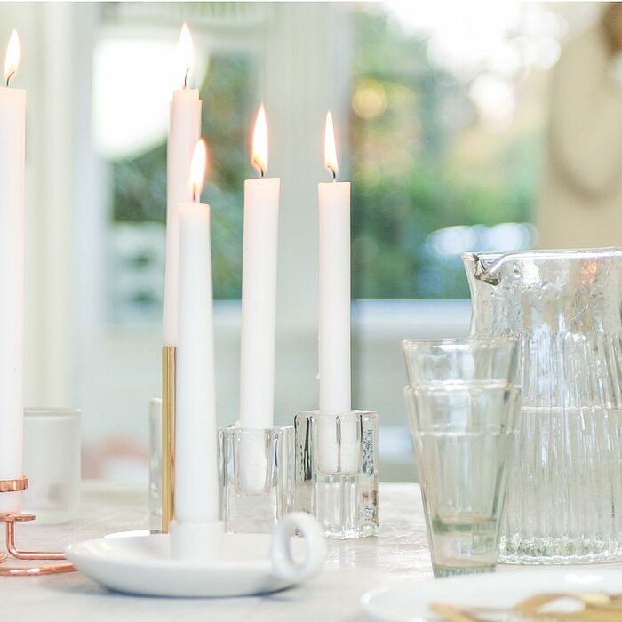 Vier brennende Bolsius Tafelkerzen in Weiß stehen in eleganten Kerzenhaltern aus Metall und Glas auf einem Tisch. Neben den Kerzen sind ein Glas-Wasserkrug und zwei Gläser zu sehen. Im Hintergrund ist eine verschwommene Szene mit einer Person und grüner U