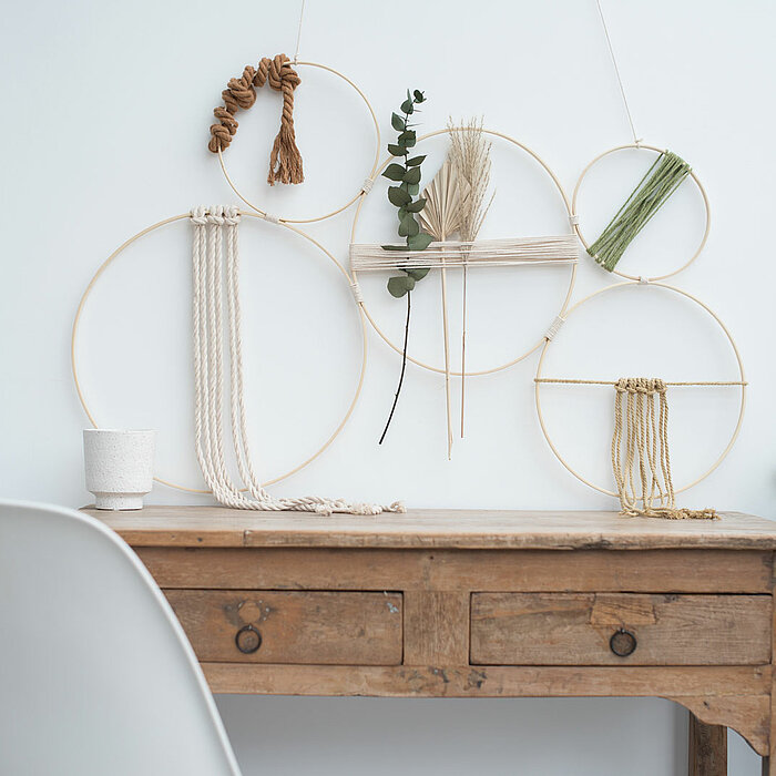 An einer weißen Wand hängen mehrere kreisförmige Holzrahmen in verschiedenen Größen, die mit Faden und natürlichen Materialien geschmückt sind. Auf einem rustikalen Holztisch darunter steht ein kleiner Becher, und links davon ist ein weißer Stuhl zu sehen