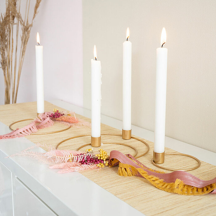 Vier brennende, weiße Stabkerzen des Herstellers "Halbach" in minimalistischen, goldenen Kerzenhaltern stehen auf einem Sideboard. Die Kerzen sind auf einem hellen Tischläufer platziert, der mit rosafarbenen und cremefarbenen Bändern sowie getrockneten Bl