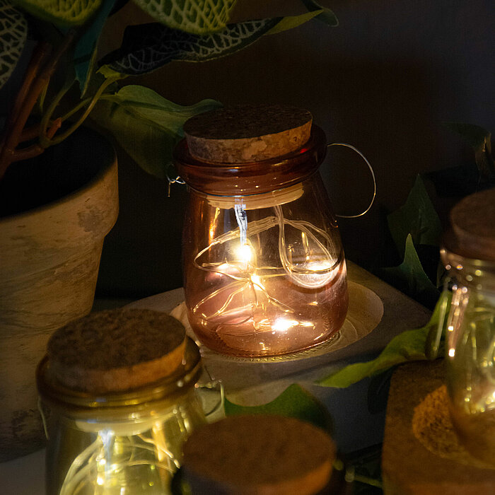 Vier Edelman Lichtergläser in verschiedenen Farben strahlen ein warmes Licht aus, verbreitet durch kleine LED-Lichterketten im Inneren. Die Gläser sind mit Korkdeckeln verschlossen und haben Henkel zum Tragen. Sie sind auf einem Tisch arrangiert, umgeben 