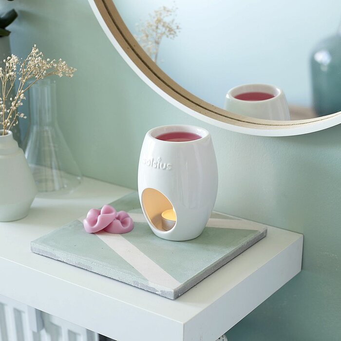Eine Bolsius-Duftkerze brennt in einem weißen Kerzenhalter mit einem offenen Design, durch den der Kerzenschein sichtbar ist. Auf dem Holztisch daneben liegen rosa Blüten und ein weiteres Duftkerzen-Accessoire. Im Hintergrund sitzt eine Person gemütlich i