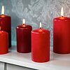 Fünf brennende rote Stumpenkerzen von DecoLite in verschiedenen Größen stehen auf einem weißen Regal. Der Hintergrund zeigt eine graue, floral gemusterte Tapete, die eine elegante Atmosphäre schafft.
