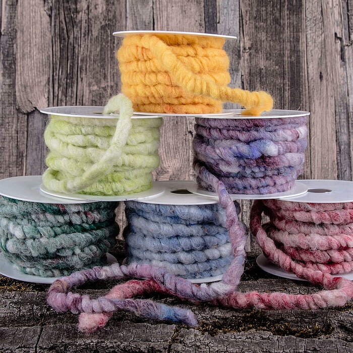 Fünf Rollen Krago Kranzbänder in verschiedenen Farben sind zu sehen, darunter Gelb, Hellgrün, Violett, Grün und Rot. Die dicken, wollartigen Bänder sind auf weißen Spulen aufgewickelt. Im Hintergrund ist eine Holzvertäfelung und der Untergrund besteht aus